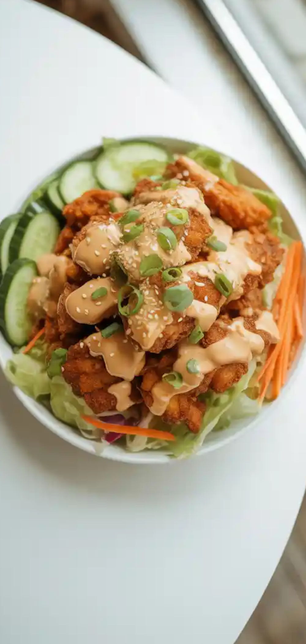 Crispy golden Bang Bang Chicken pieces drizzled with creamy spicy sauce served with fresh shredded carrots, cucumber slices, and lettuce in a white bowl on a bright white table.