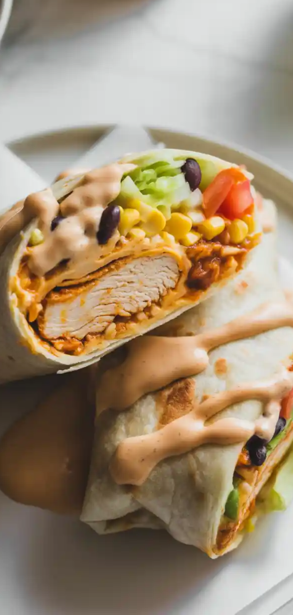 “Overhead shot of a colorful grilled chicken burrito with chipotle ranch sauce, served with corn and diced tomatoes, styled on a clean white table with natural lighting.”