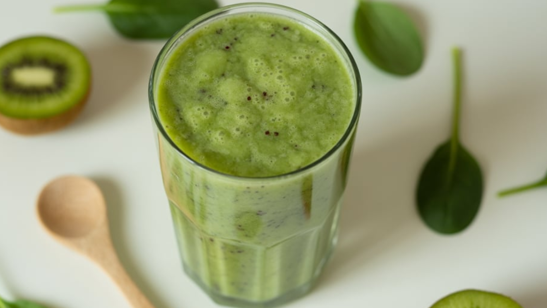 A refreshing Kiwi Spinach Smoothie in a tall glass on a white table with fresh kiwi slices and spinach leaves around.
