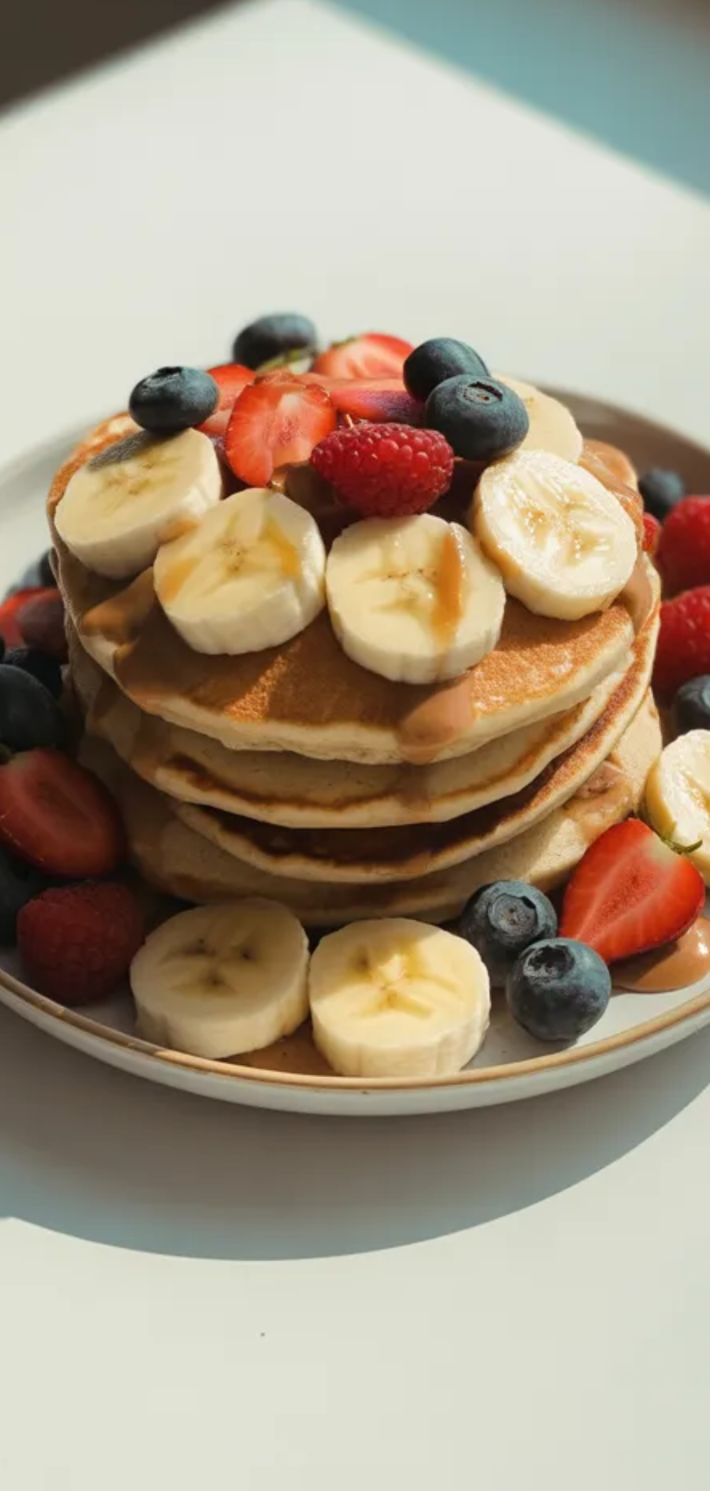 Protein-packed breakfast with pancakes, banana slices, and berries on a bright white table, natural lighting, fresh and healthy presentation.