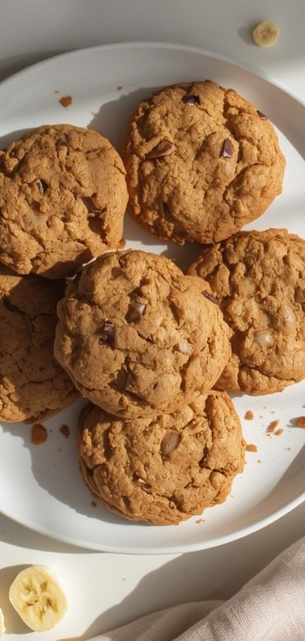 “Homemade soft and chewy Banana Oat Cookies arranged neatly on a plate, crumbs and chocolate chips scattered, bright food photography style”