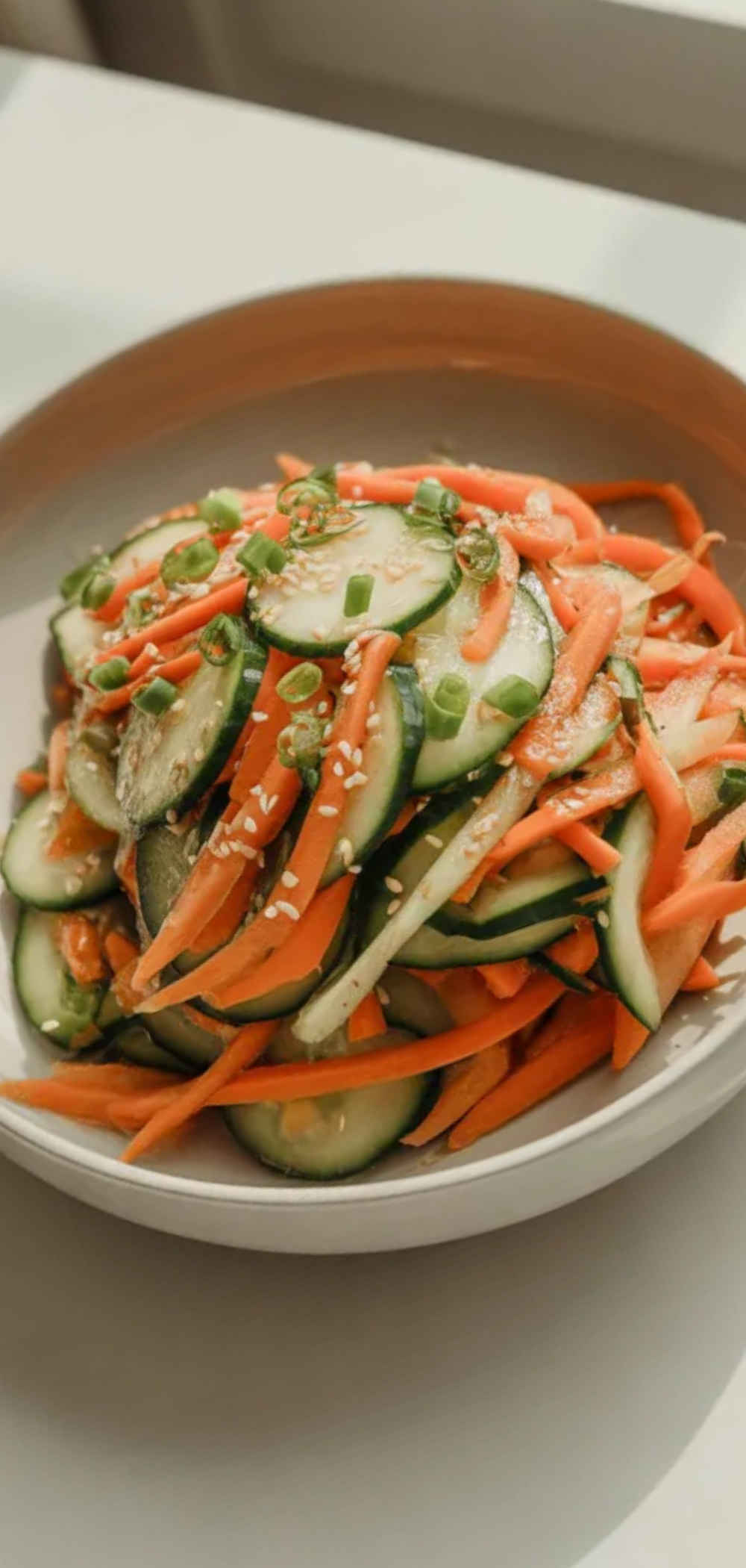 Top view of Asian carrot and cucumber salad in a white bowl on a clean white table with sesame seeds and green onions