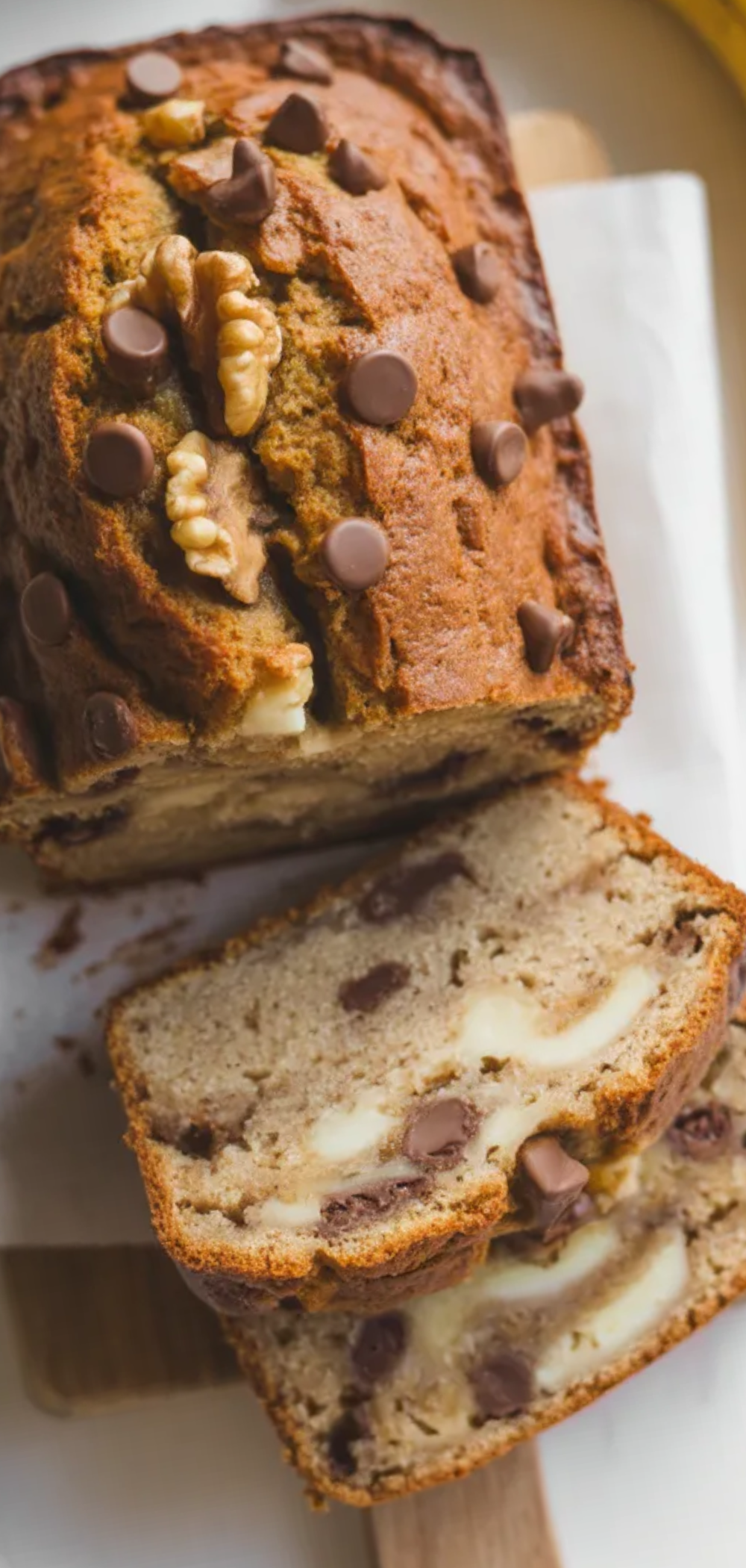 top down of a freshly baked banana bread loaf made with cottage cheese, sliced to show its soft, moist interior, styled on a white table with natural light, chocolate chips and walnuts scattered on top.