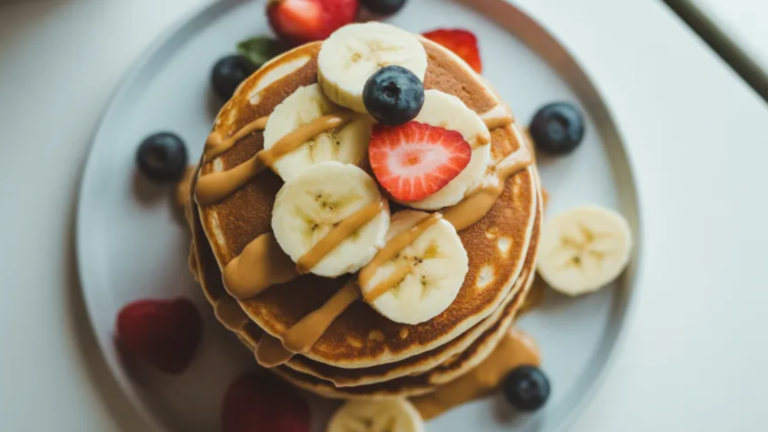 Golden protein pancakes with fresh banana and berries on a white table, styled with natural light, wholesome and kid-friendly breakfast.