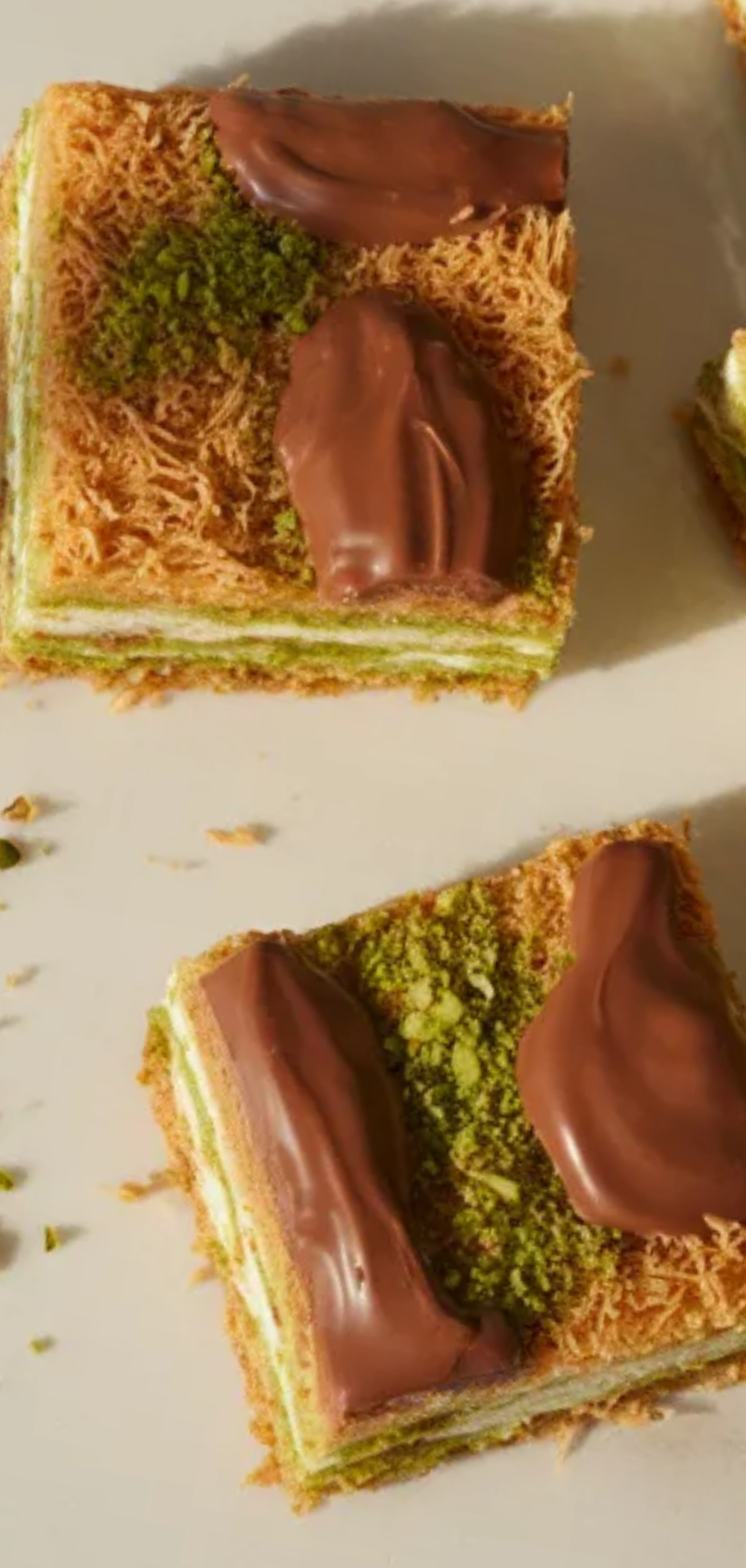 A plated dessert of pistachio kunafa chocolate bars with visible crunchy kunafa base, creamy pistachio layer, and smooth chocolate coating, garnished with crushed pistachios.