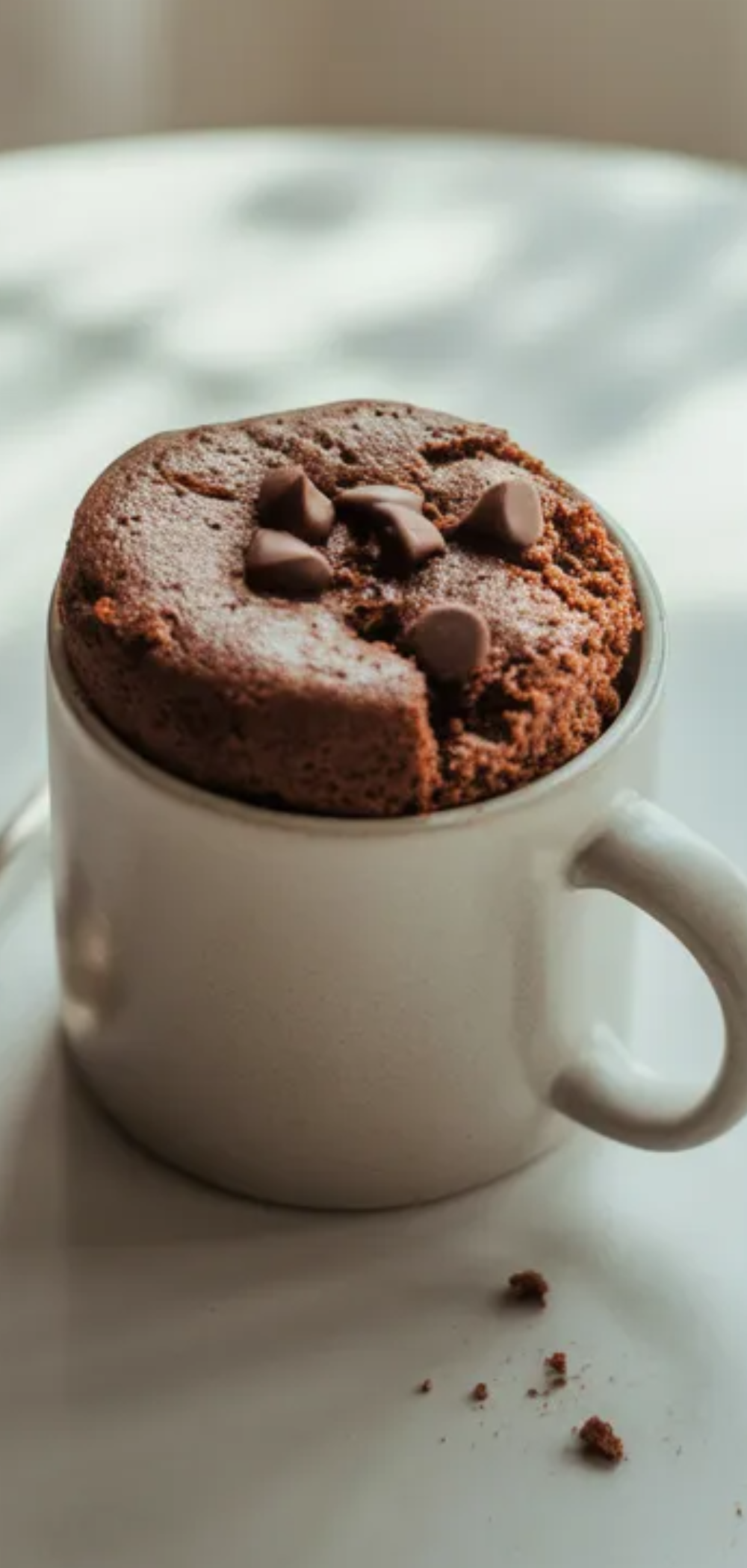 Minimalist food photography of a warm mug cake on a white surface with soft daylight and a cozy homemade dessert style.