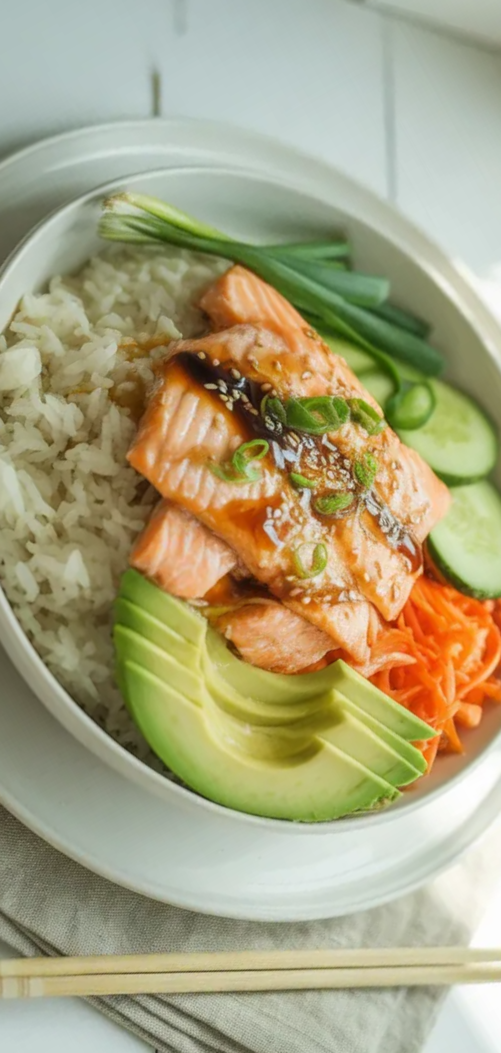 Top view of homemade salmon rice bowl with glazed salmon, rice, fresh vegetables, and soy sauce drizzle on a bright minimal white background