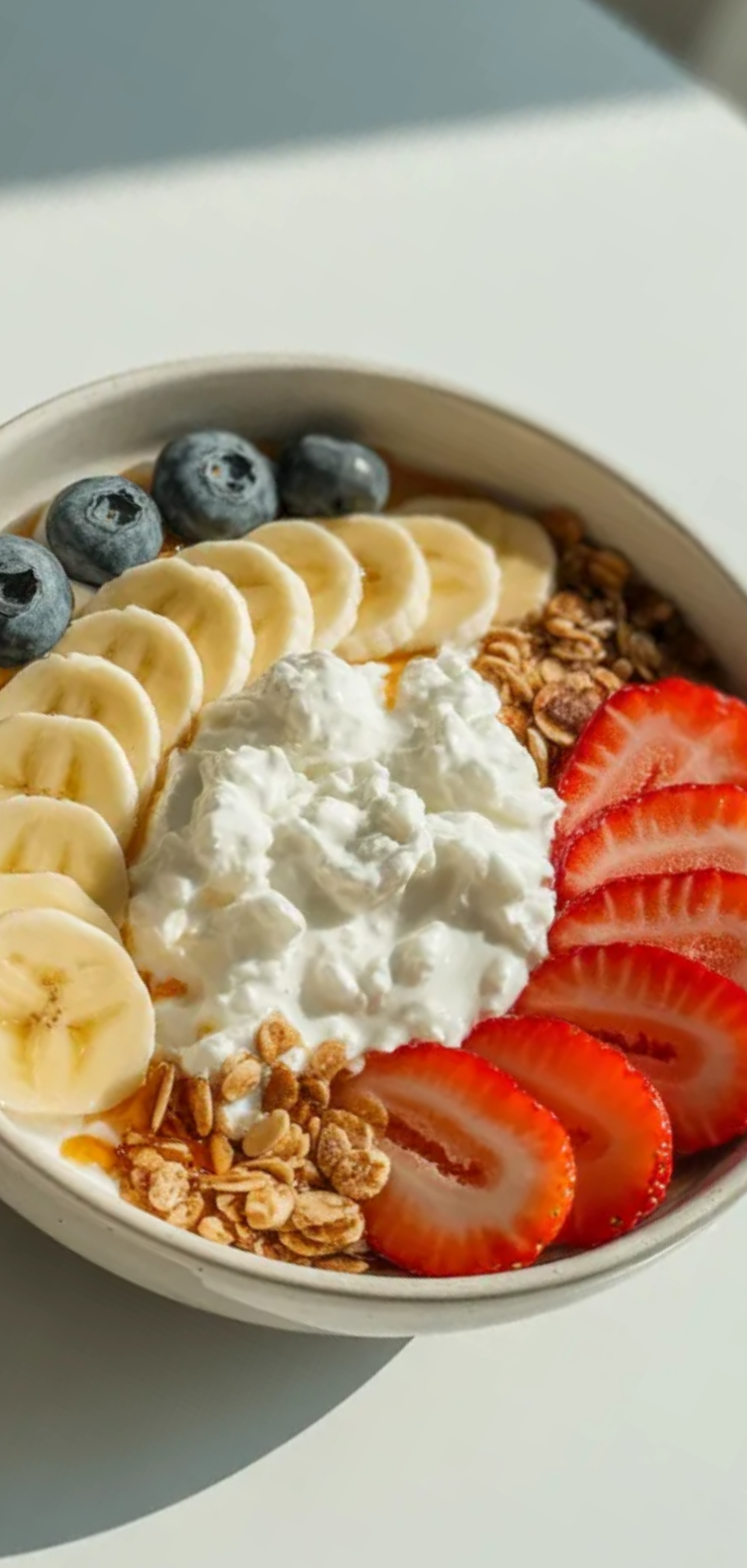 Top-down view of a healthy cottage cheese bowl with mixed berries, banana slices, and granola, presented as a quick high-protein breakfast on a bright white table.

