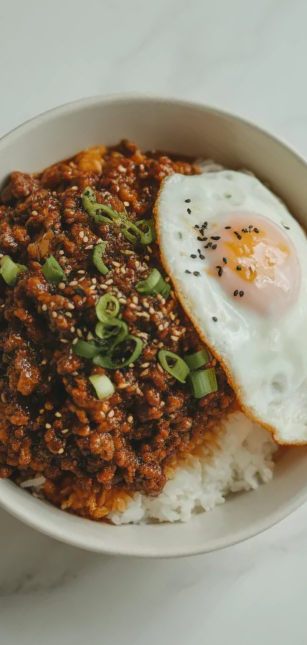 Top-down view of a homemade Korean Ground Beef Bowl in a white dish, featuring savory beef over rice with a soft fried egg on top, minimal styling and bright natural kitchen lighting.