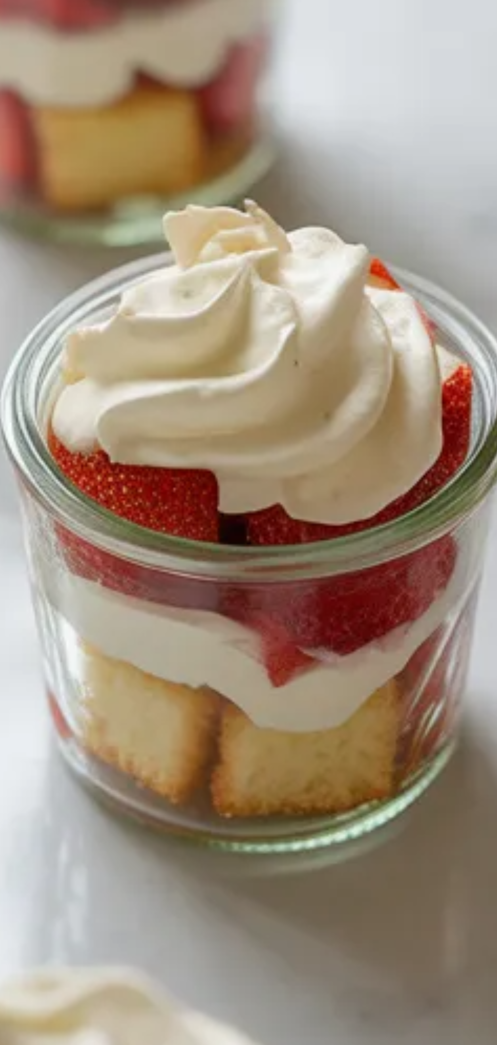 Homemade strawberry dessert in jars with visible layers of cake, whipped cream, and fresh strawberries styled on a bright minimal background.