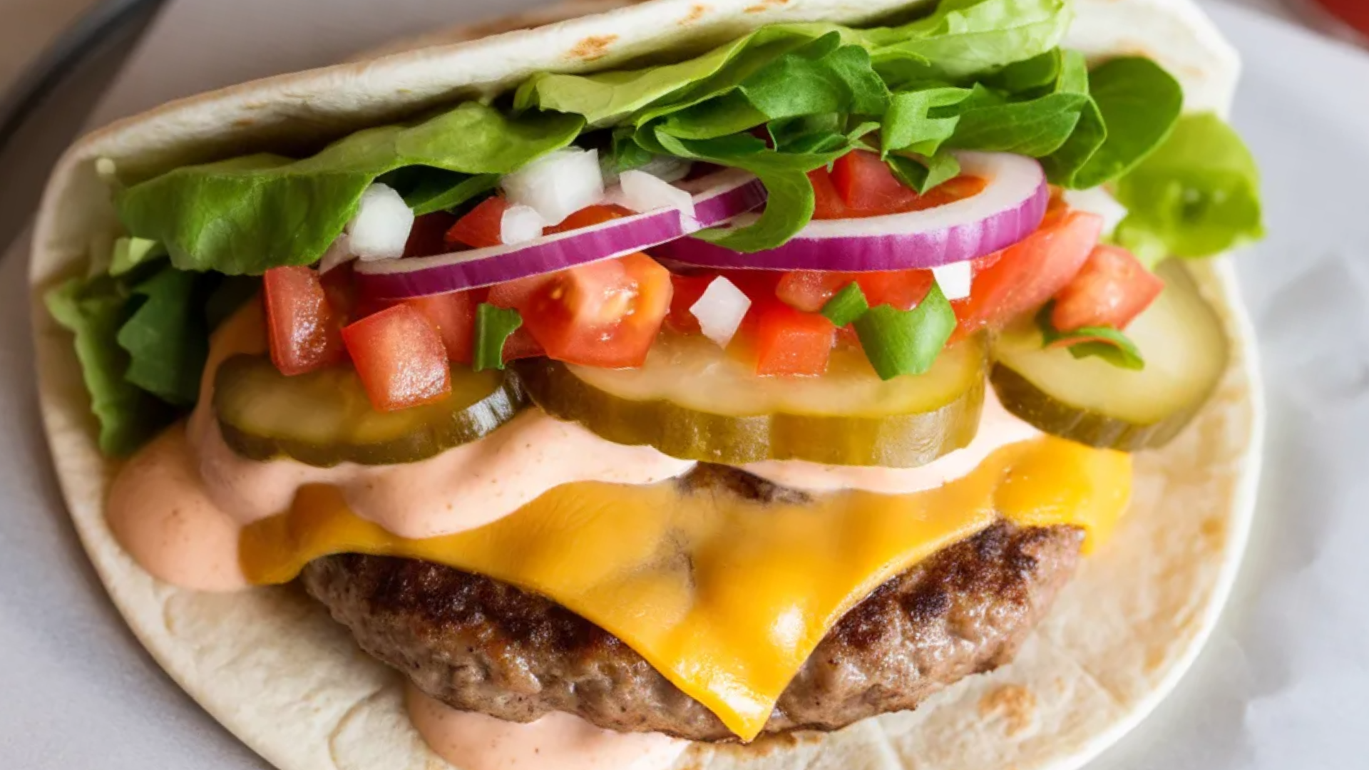 “Close-up of a Smash Burger Taco with melted cheddar, lettuce, tomato, onions, and pickles on a soft flour tortilla on a white table.”