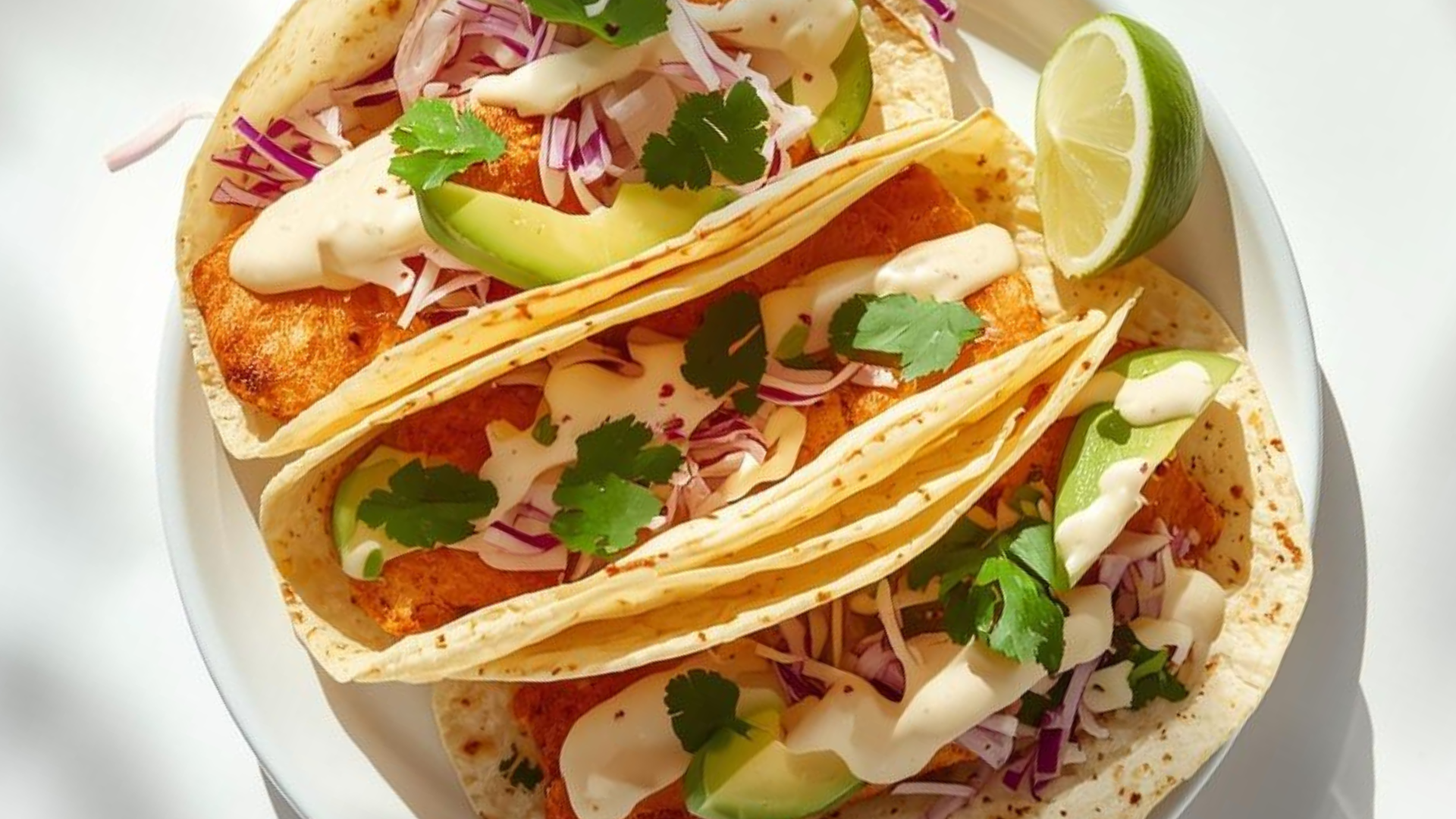 A plate of freshly made fish tacos with crispy white fish, shredded cabbage, avocado slices, and creamy sauce on a white table in natural daylight.