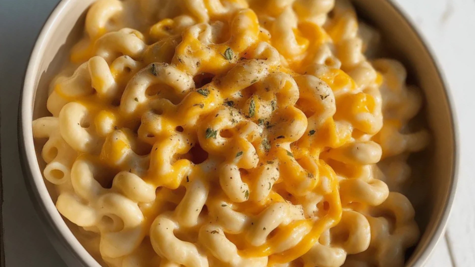 “A white bowl filled with creamy mac and cheese topped with melted cheddar and mozzarella, sitting on a white wooden table with soft natural lighting.”