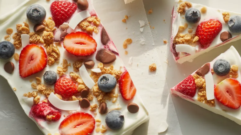 Frozen yogurt bark broken into pieces on a white marble table topped with strawberries, blueberries, chocolate chips, and granola.
