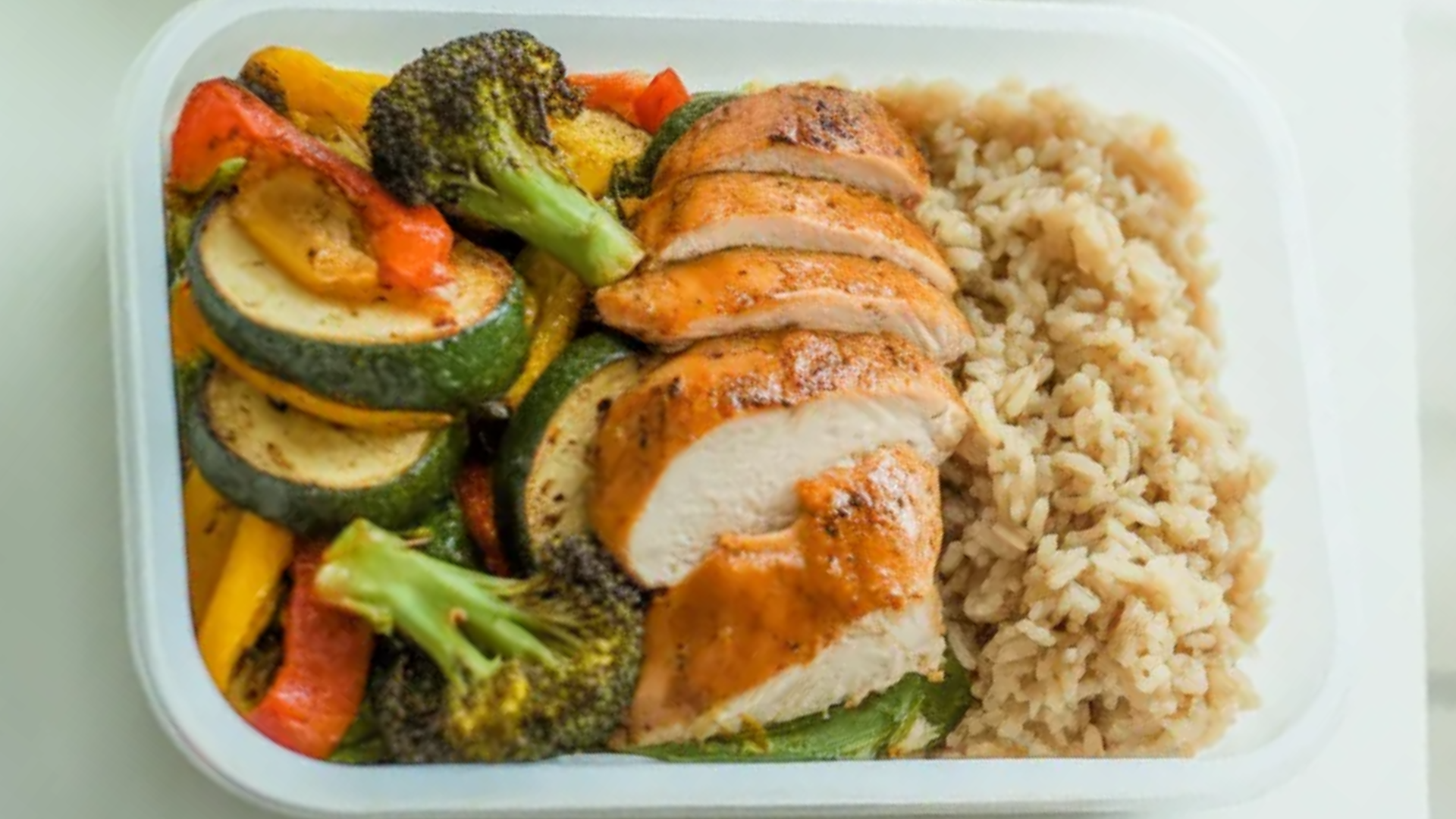 Healthy chicken meal prep with grilled chicken breast, roasted vegetables, and brown rice in white containers on a clean white table with soft natural lighting