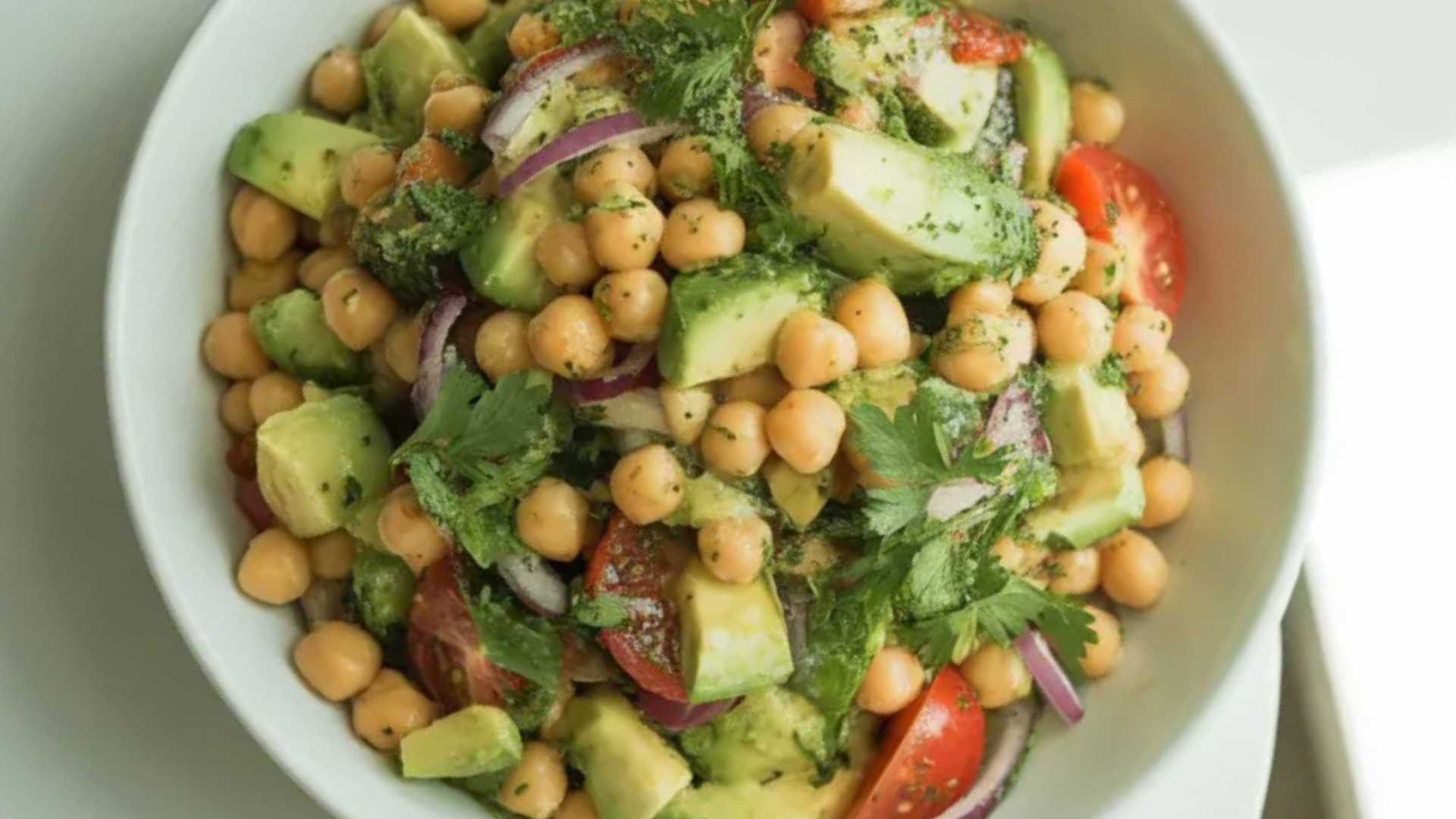 Chickpea avocado salad in a white bowl with fresh vegetables and lemon dressing on a white table