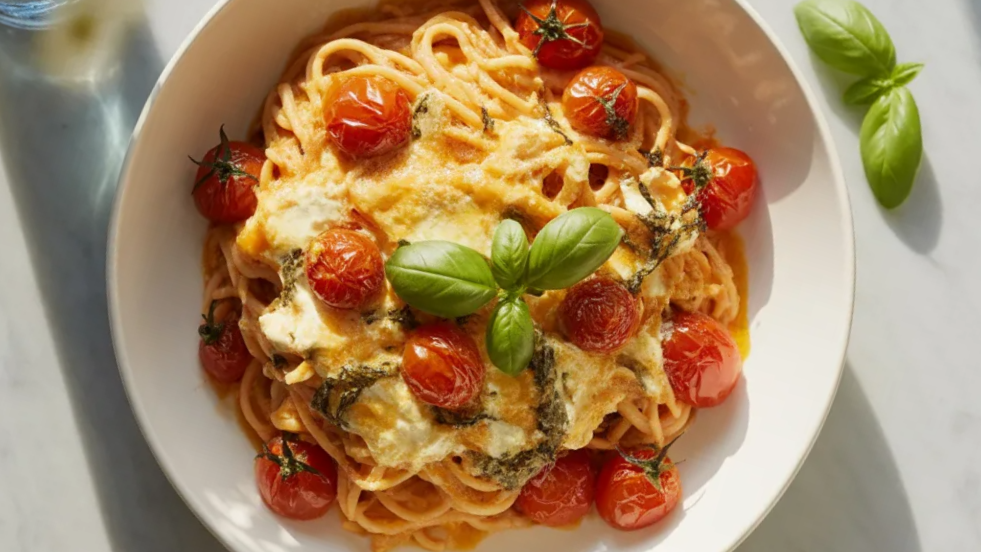Creamy baked feta pasta served in a white bowl with roasted cherry tomatoes, melted feta cheese, and fresh basil on top on a white marble table.