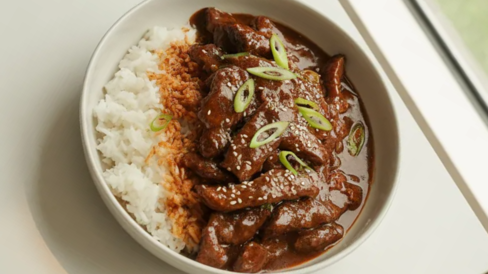 Homemade Mongolian Beef stir fry with tender beef strips, rich soy garlic sauce, and steamed rice on a white table in natural light.