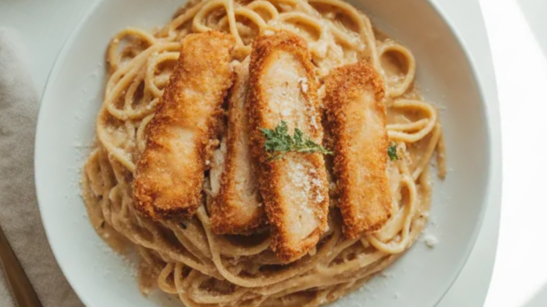 Crispy golden chicken strips served over creamy garlic pasta on a white plate, styled as a cozy homemade comfort food dish.