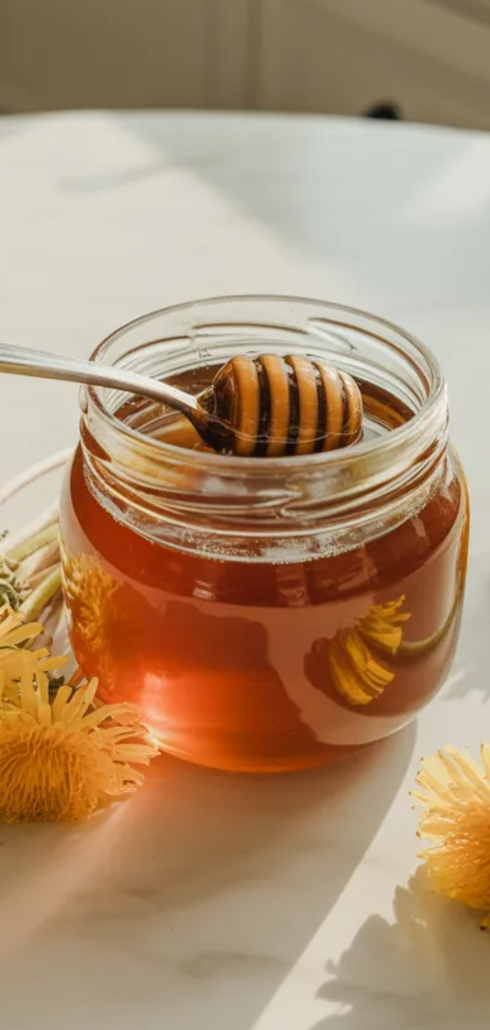 Homemade vegan dandelion honey in a glass jar with a spoon inside, styled on a bright white table with soft natural light and minimalist Pinterest food blog design text in the middle.