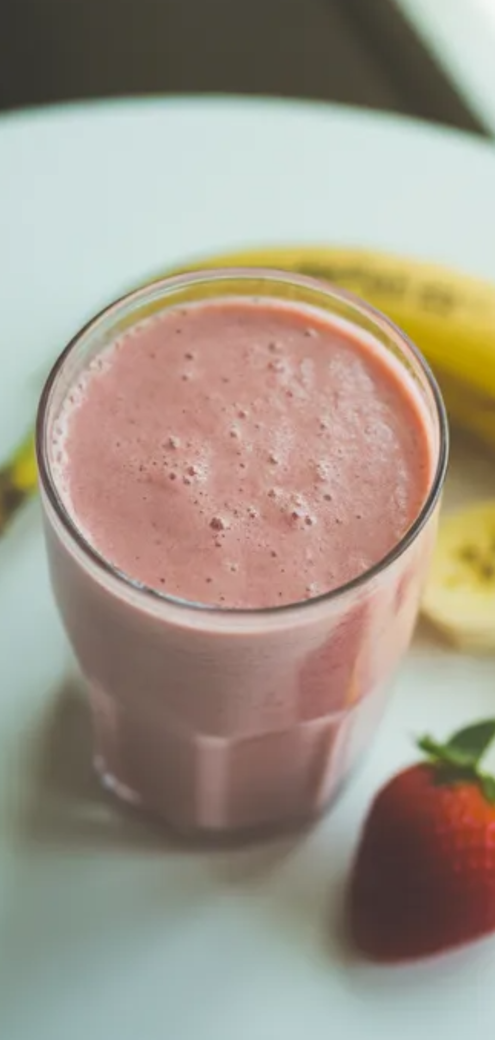 Fresh strawberry banana smoothie in a minimalist white kitchen setting, creamy pink color, served cold with fruit garnish and soft daylight lighting.